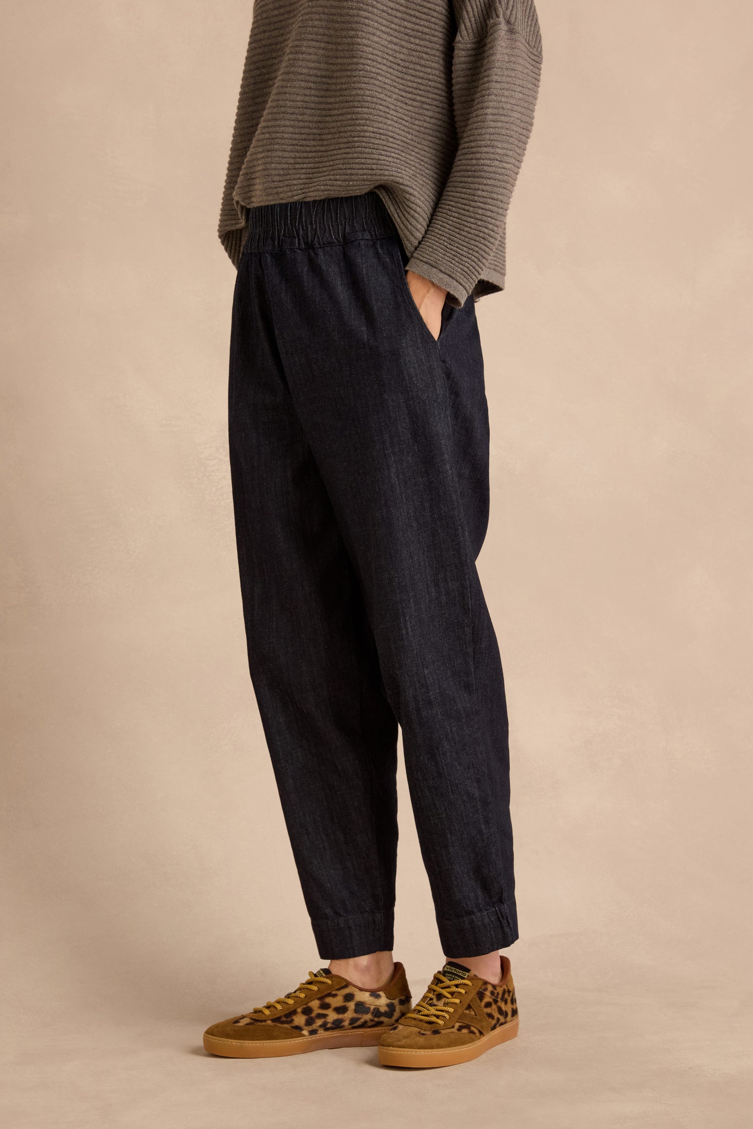 Wearing a gray ribbed sweater, Laura Tapered Denim Trousers made from pure cotton with an elasticated waistband and pockets, and brown leopard print sneakers, the person stands against a beige background.