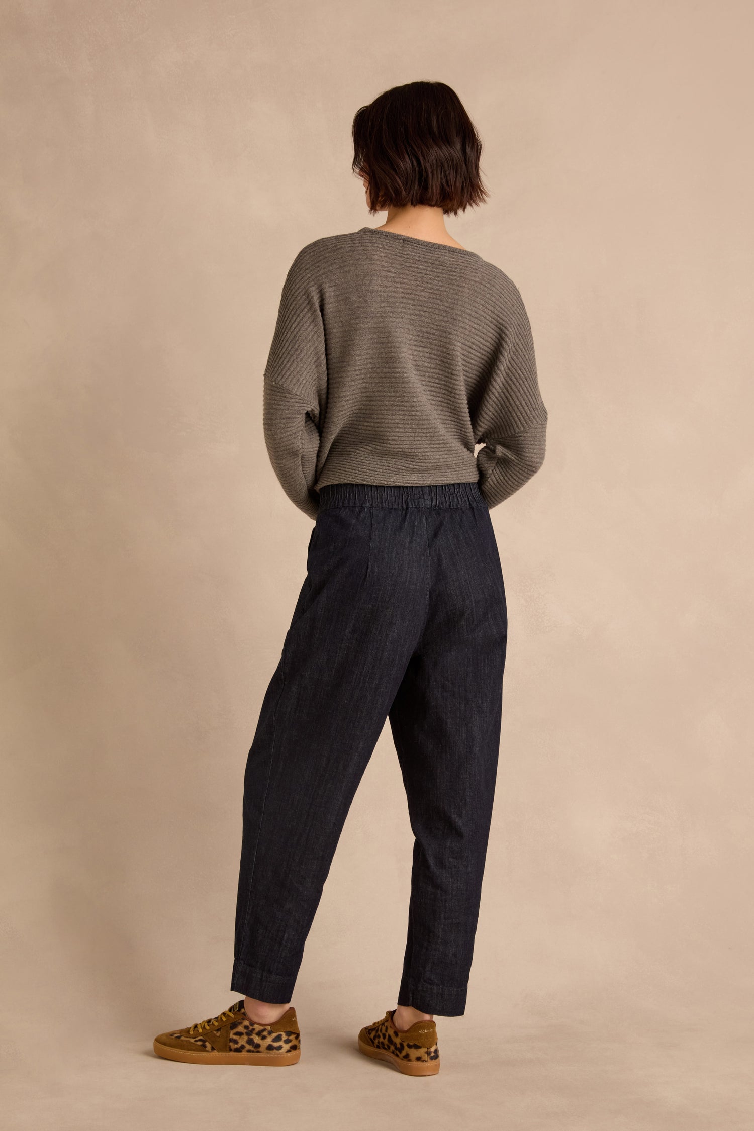 A person stands facing away against a beige background, wearing a gray sweater, Laura Tapered Denim Trousers with an elasticated waistband made from pure cotton denim, and leopard-print shoes.