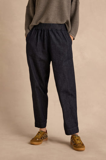 A person wearing a loose grey sweater, the Laura Tapered Denim Trousers in dark blue pure cotton with a high-waisted elasticated waistband, and leopard print sneakers, stands with hands in pockets against a beige background.
