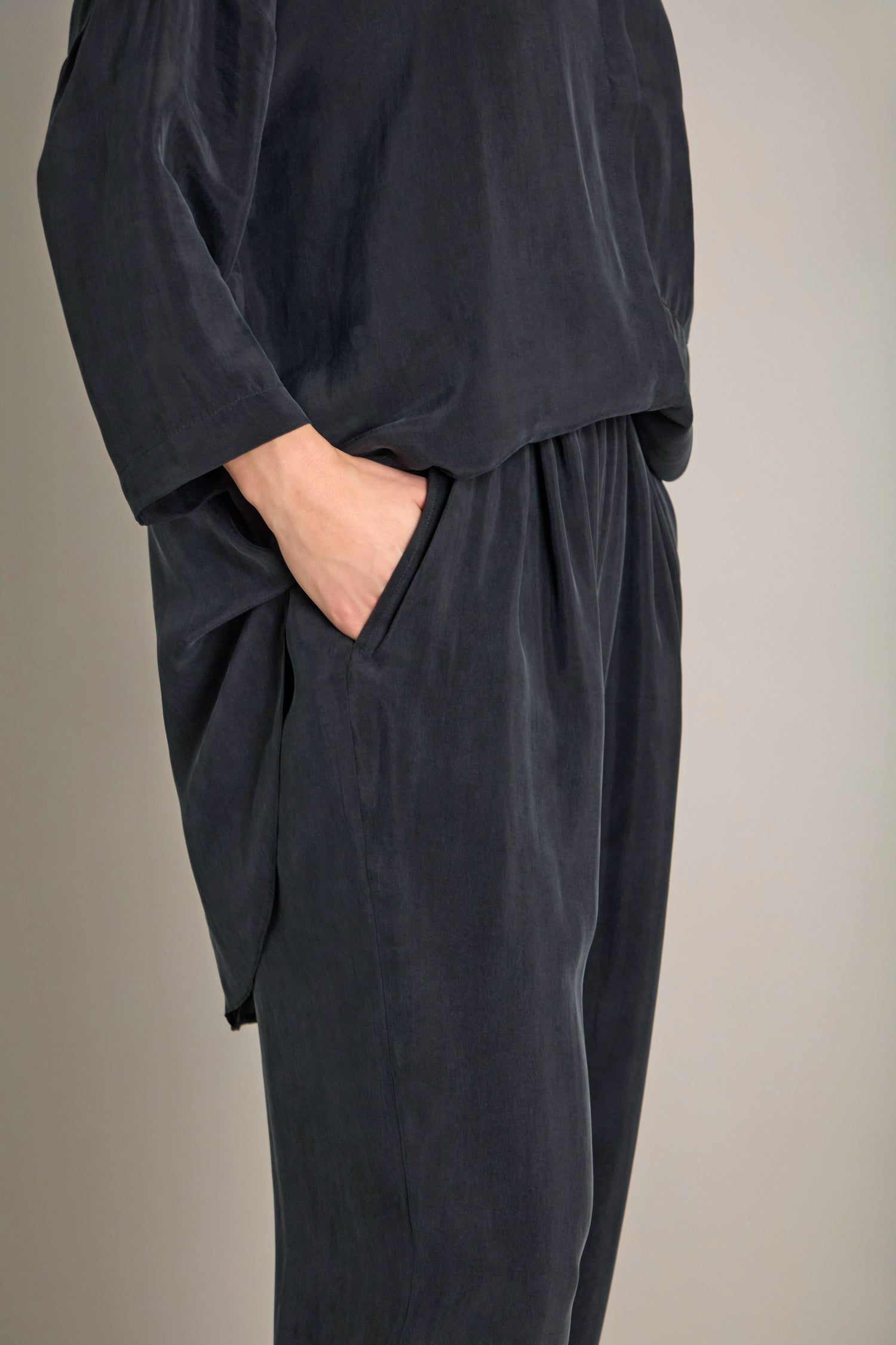 A person elegantly dressed in a loose-fitting dark gray outfit with one hand in their pocket, showcasing the Lola Drapey Cupro Trouser—an essential piece for any modern wardrobe with its elasticated waistband.