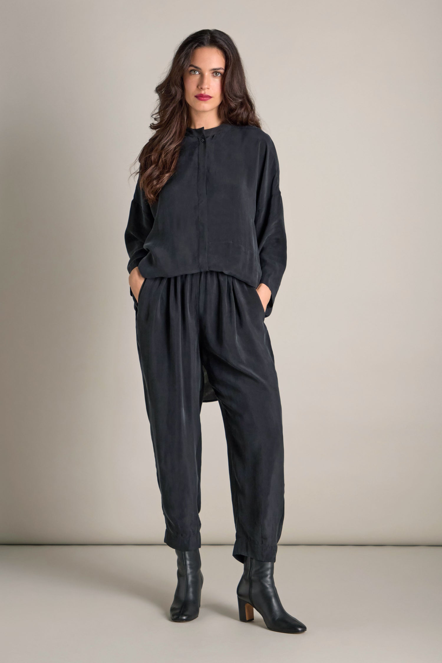 Dressed in a relaxed black ensemble, which includes the Lola Drapey Cupro Trouser with its comfortable elasticated waistband and black ankle boots, an individual stands against a simple backdrop.