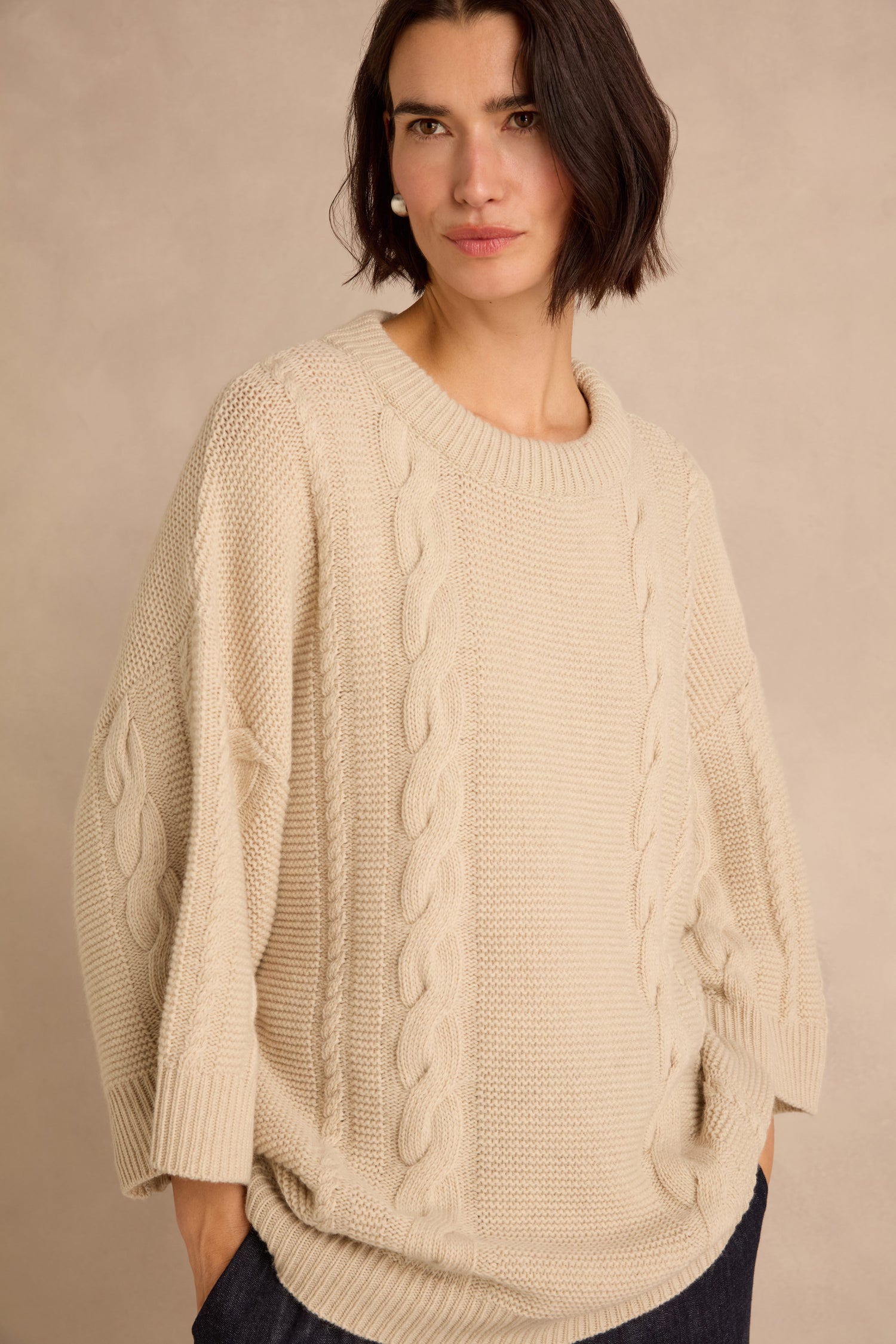 A woman stands against a plain beige background with her hands in the pockets of a loose, cream-colored Cable Detail Wool Tunic made from recycled wool.