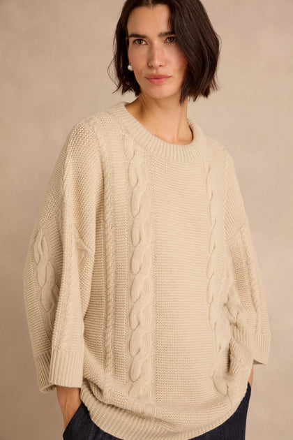A woman stands against a plain beige background with her hands in the pockets of a loose, cream-colored Cable Detail Wool Tunic made from recycled wool.