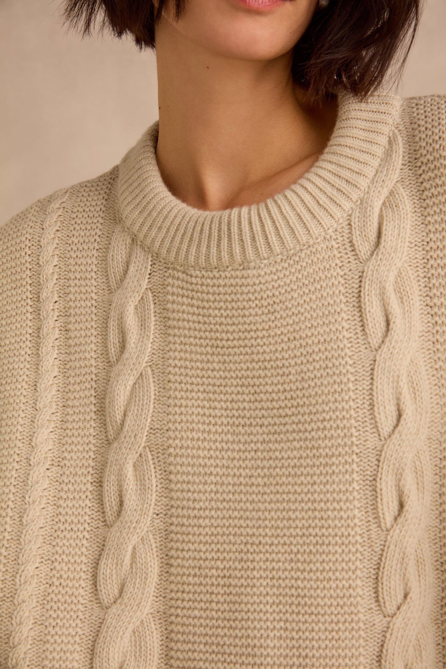 A person wearing the Cable Detail Wool Tunic in beige, featuring a ribbed collar and crafted from recycled wool. Only the lower half of their face and upper torso are visible.