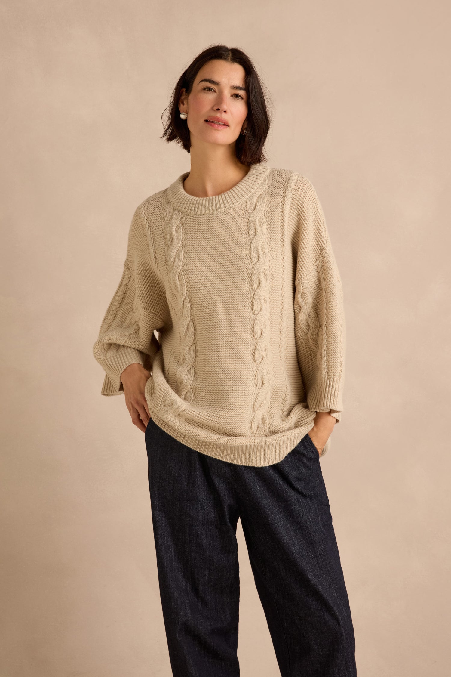 A woman stands against a plain, light-colored background wearing the Cable Detail Wool Tunic—a loose, beige cable knit piece crafted from recycled wool—paired with dark wide-leg pants.