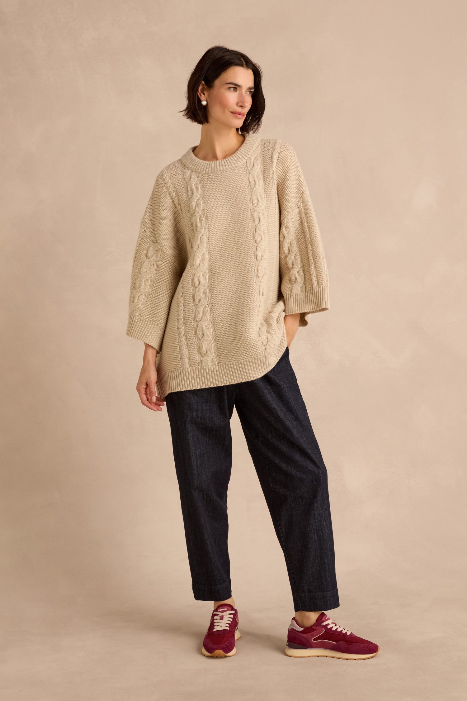 Woman wearing a Cable Detail Wool Tunic in beige, paired with dark loose-fitting pants and red sneakers, standing against a light beige background.