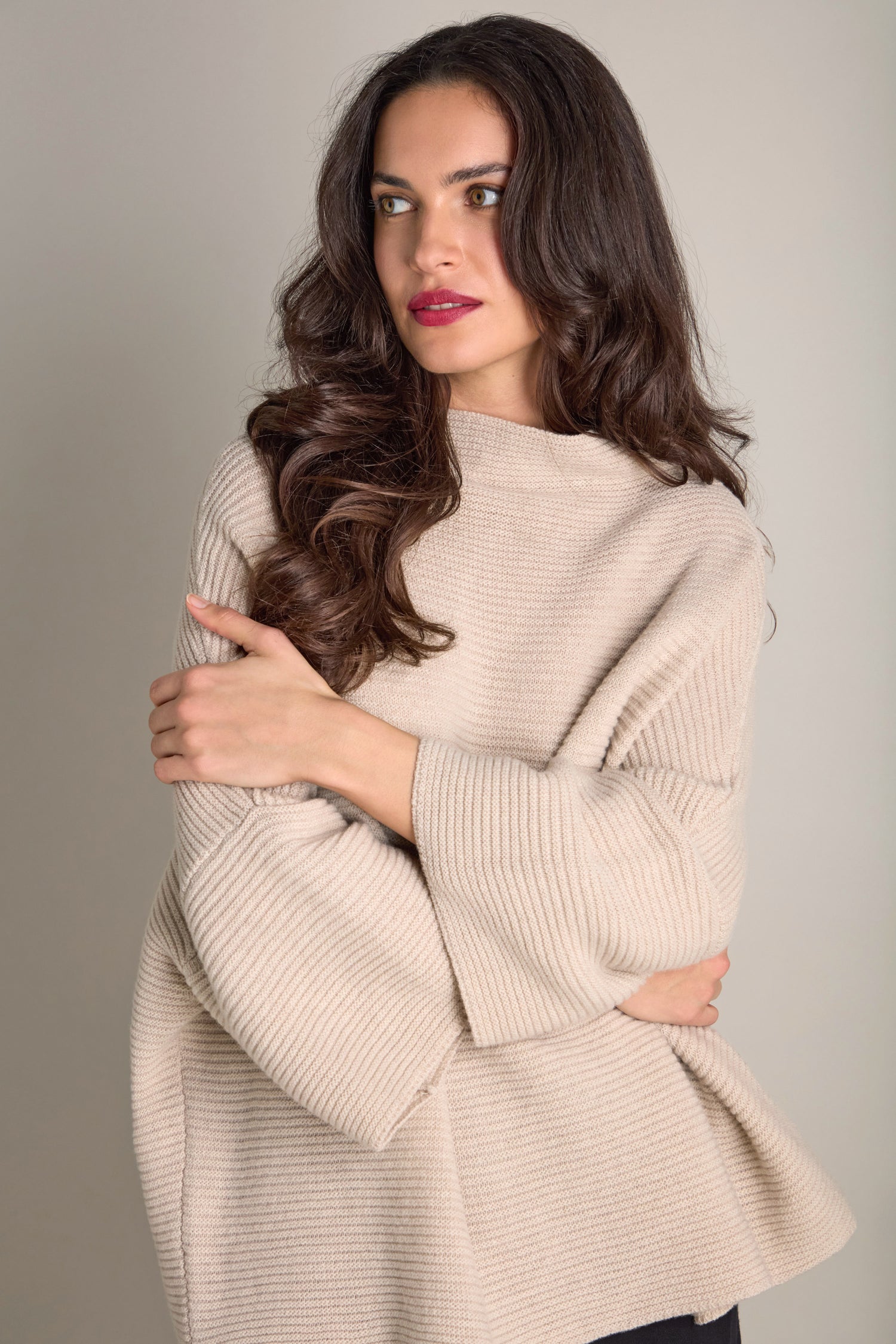 A woman with long, wavy hair wearing the Pauline Rib Boxy Knit in an oversized fit stands against a neutral background, looking to the side with her arms crossed.