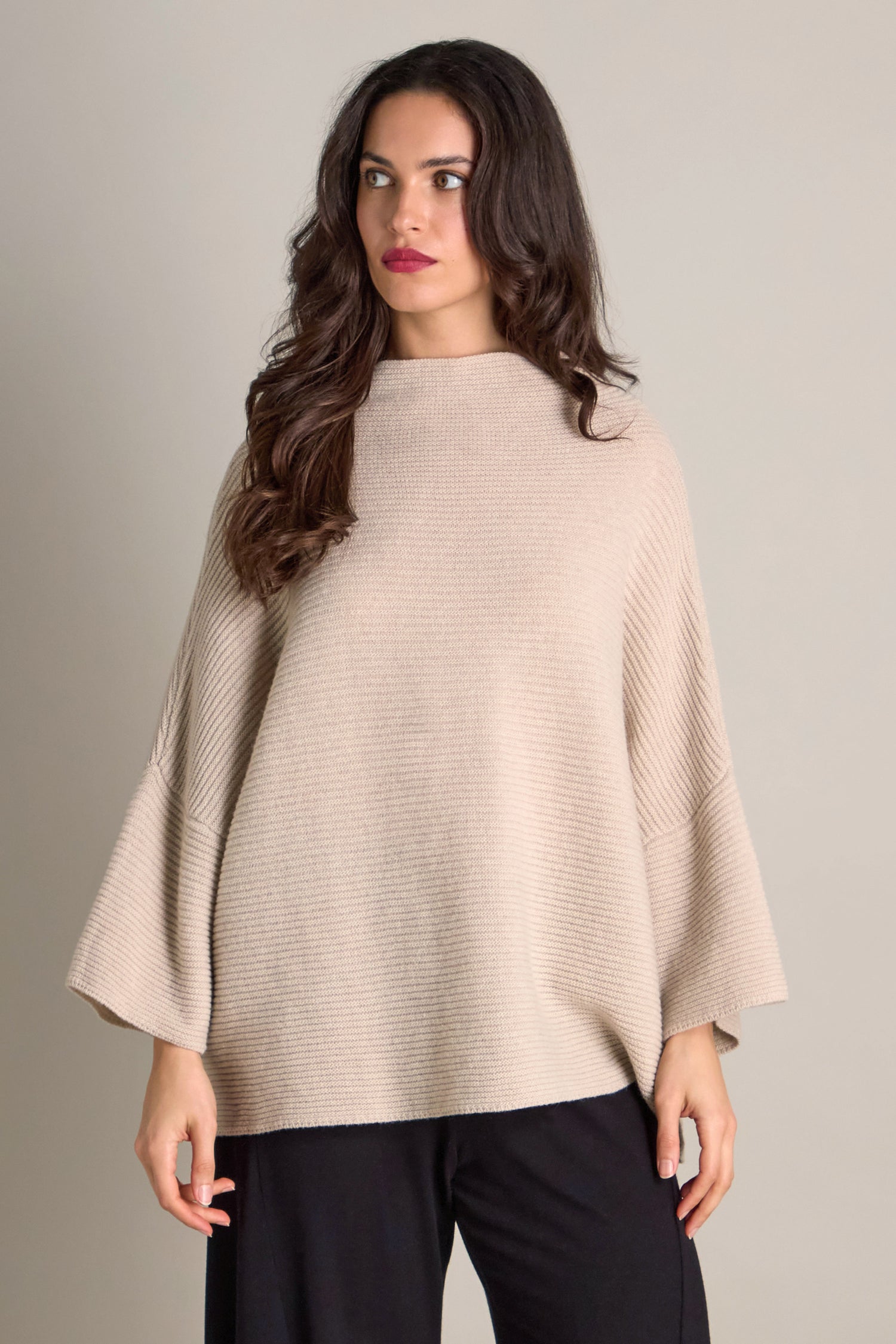 A woman with long dark hair, draped in the Pauline Rib Boxy Knit, stands against a plain background, exuding an aura of cozy comfort as she gazes to the side.