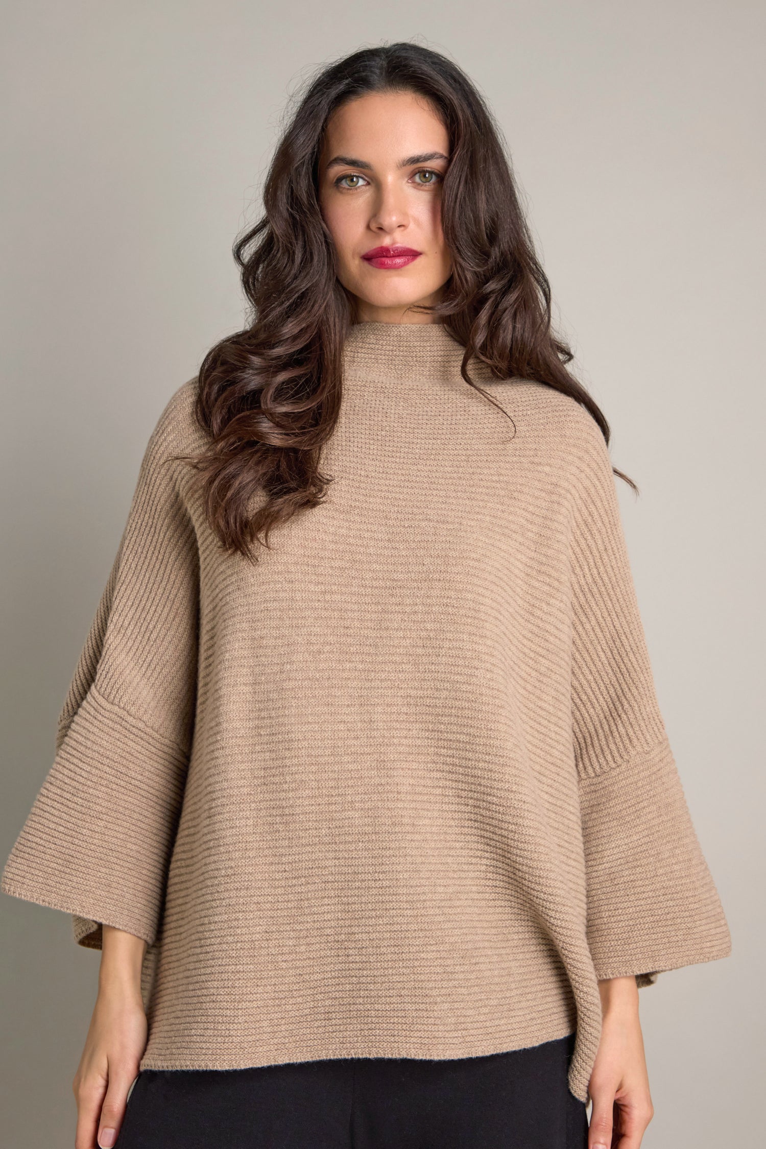 A woman with long dark hair wears the Pauline Rib Boxy Knit, an oversized beige poncho-style sweater, exuding cosy comfort against a neutral background.