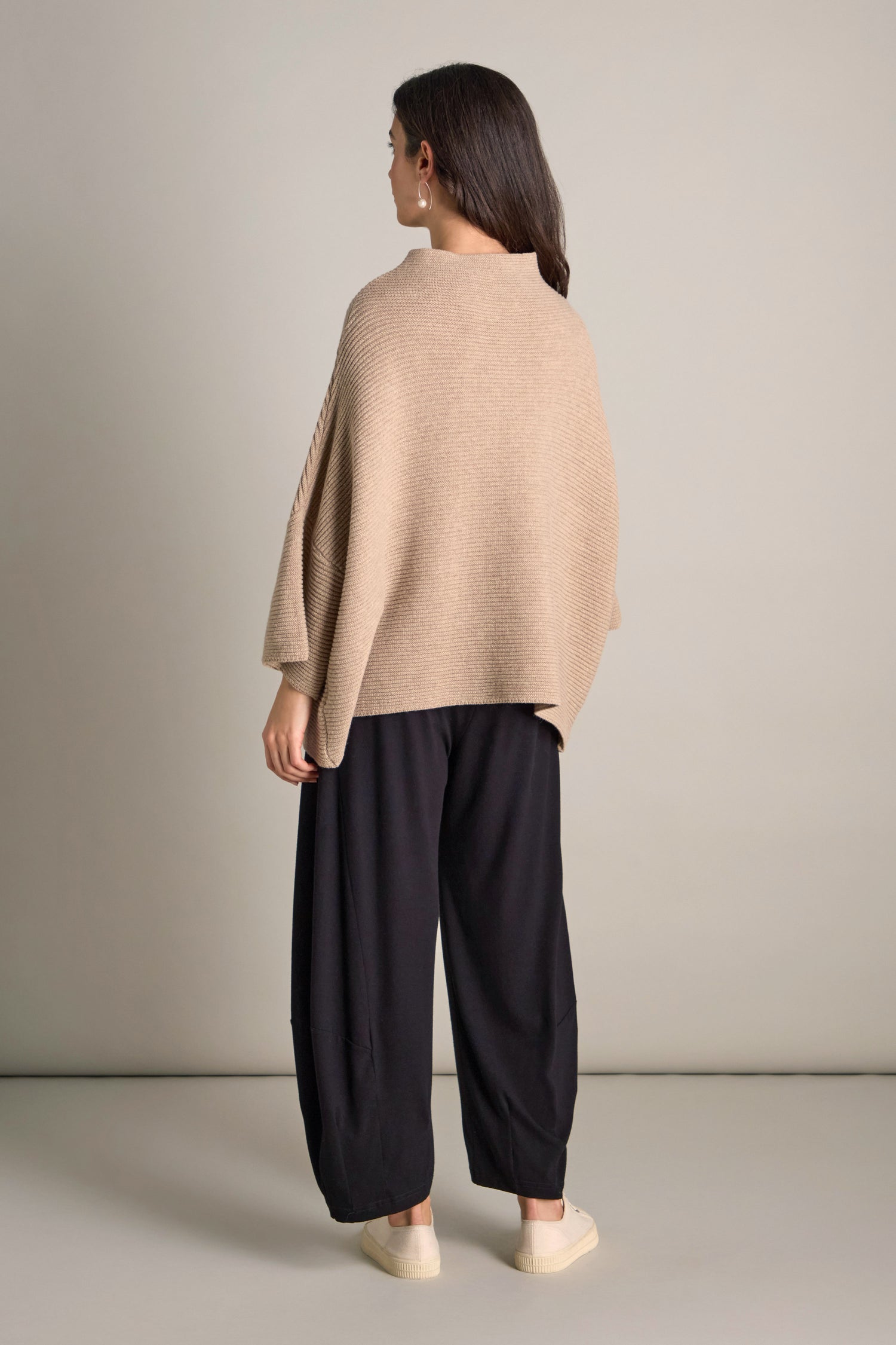 A person with long dark hair is standing sideways, wrapped in the Pauline Rib Boxy Knit and black wide-leg pants. Their cozy comfort is completed by beige shoes against a plain background.