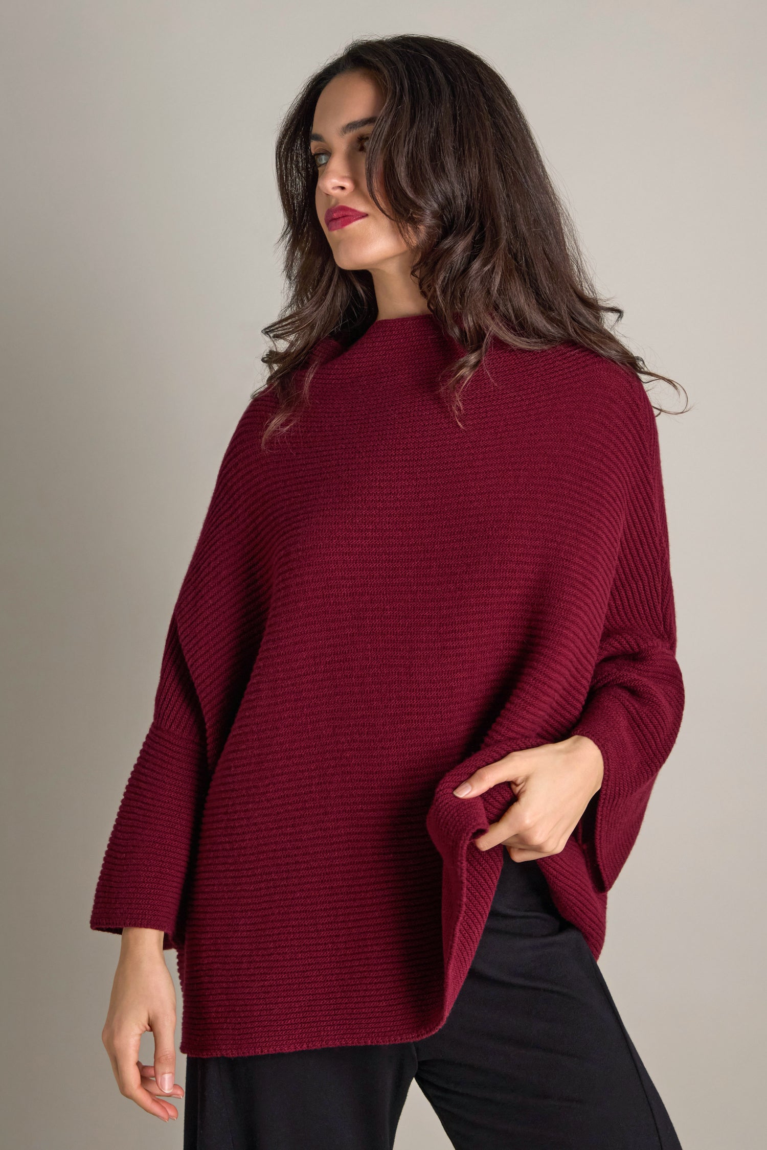 A woman dressed in the Pauline Rib Boxy Knit, an oversized dark red sweater with a textured design, alongside black pants, stands against a plain background, exuding cosy comfort.