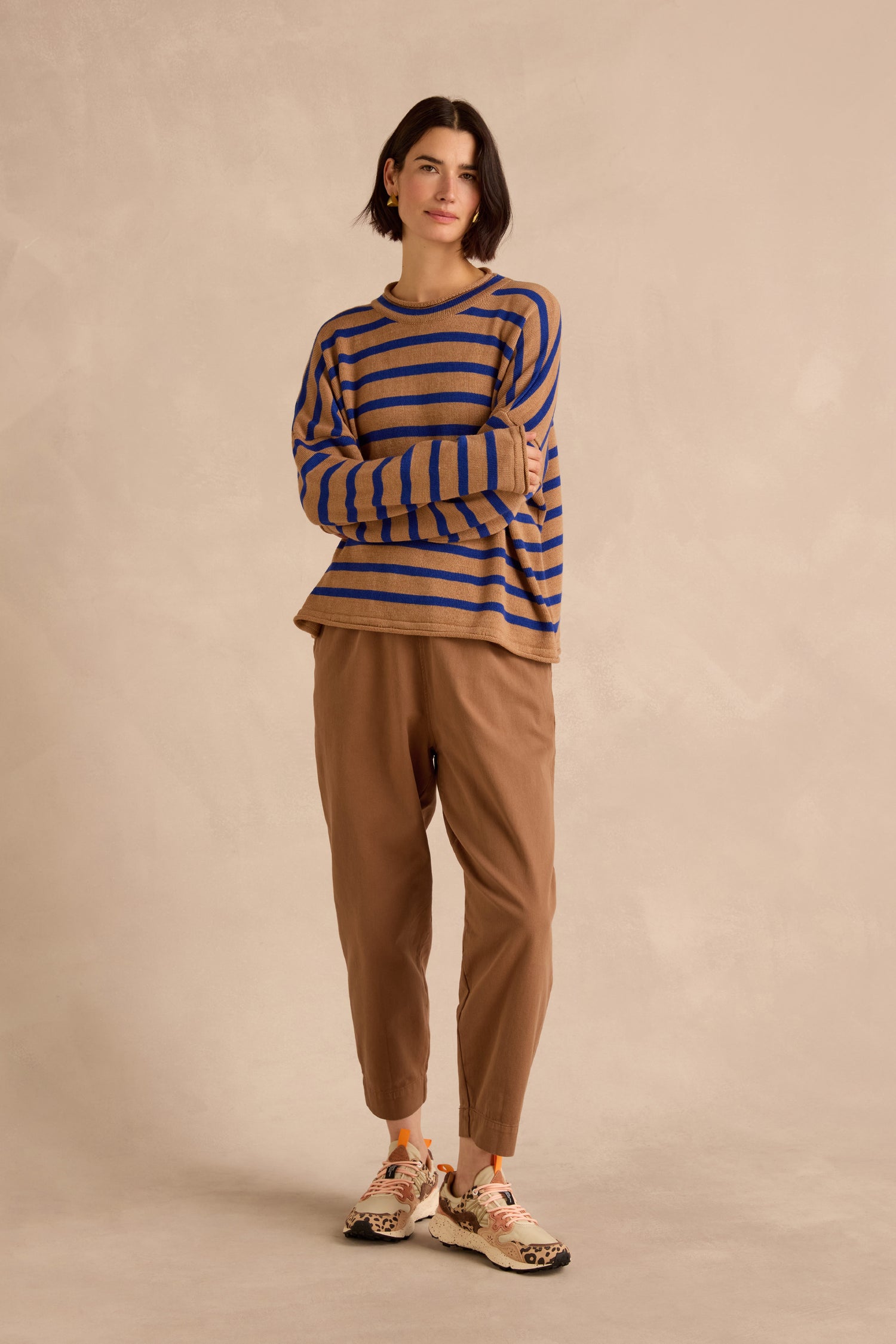 A person stands against a beige background, wearing the Royal Blue Stripe Boxy Merino Knit sweater—crafted from soft merino wool with blue and tan stripes—paired with tan pants and animal-print-accented sneakers.