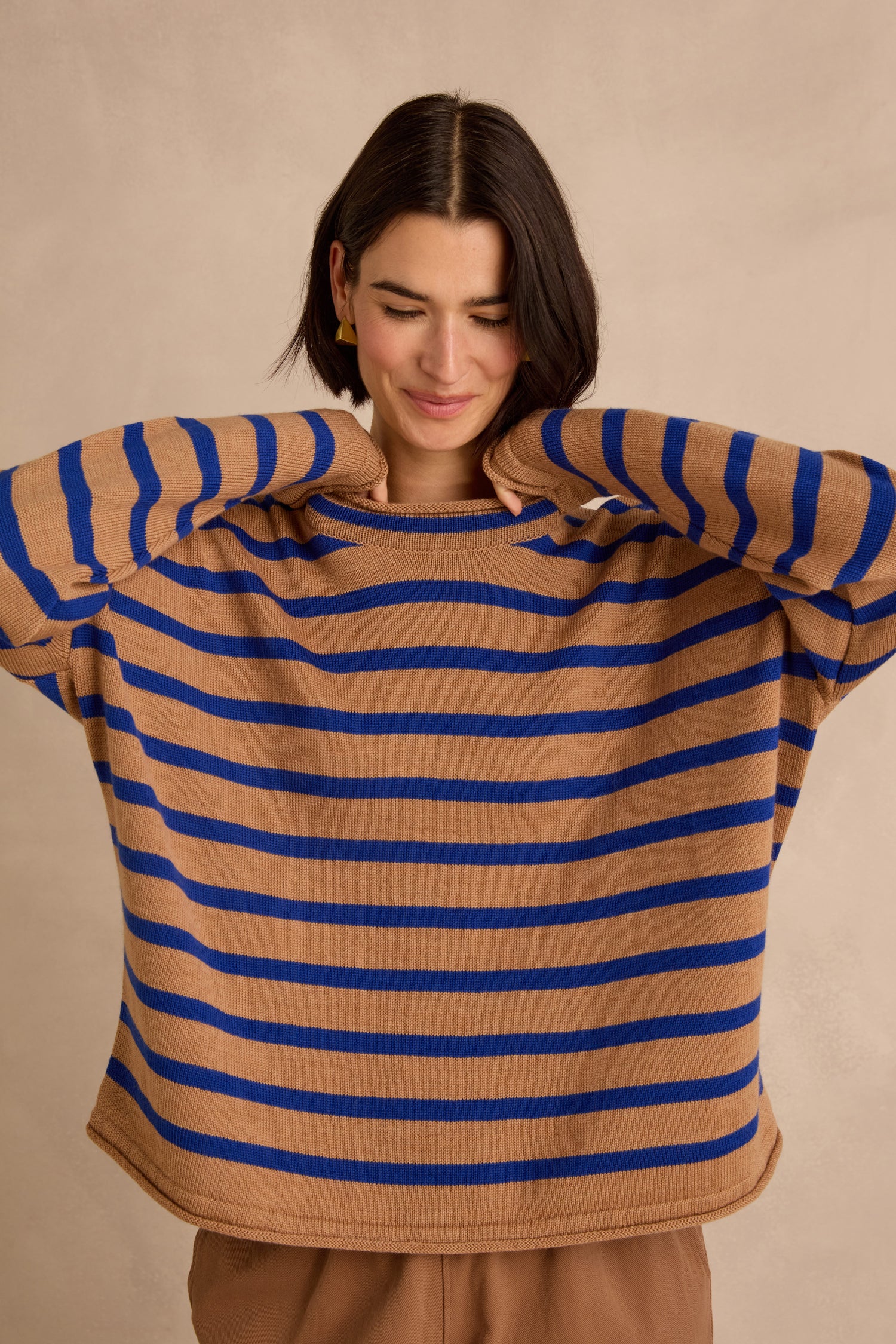 A person with short dark hair wears the Royal Blue Stripe Boxy Merino Knit—a loose, boxy brown sweater featuring bold royal blue horizontal stripes—standing against a plain beige background with arms raised and a slight smile.