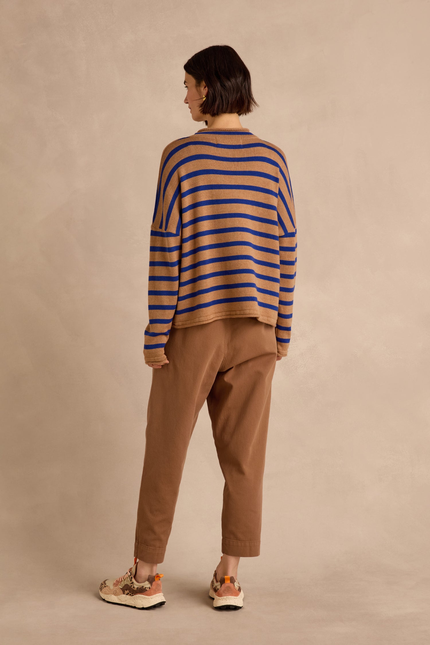 A person stands with their back to the camera, wearing the Royal Blue Stripe Boxy Merino Knit sweater—crafted from soft merino wool in blue and tan stripes—paired with brown pants and multicolored sneakers, set against a neutral beige background.