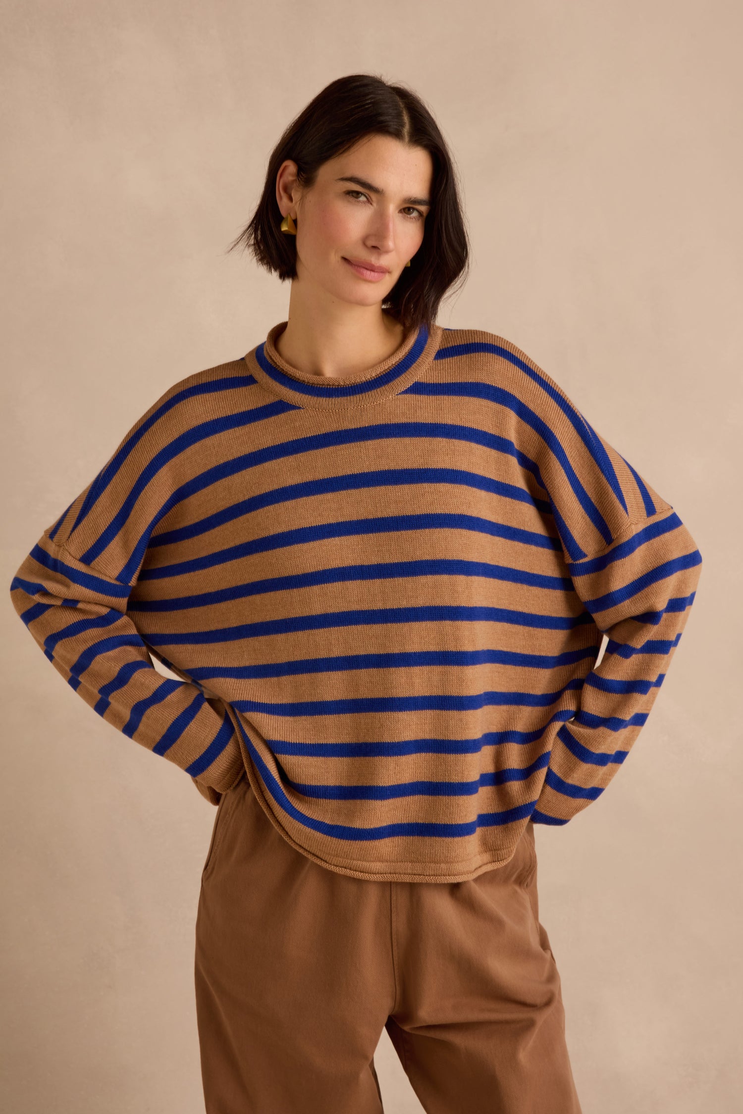 A woman with short dark hair stands with her hands on her hips against a beige background, wearing the Royal Blue Stripe Boxy Merino Knit sweater—crafted from soft merino wool in brown and blue stripes—paired with brown pants.