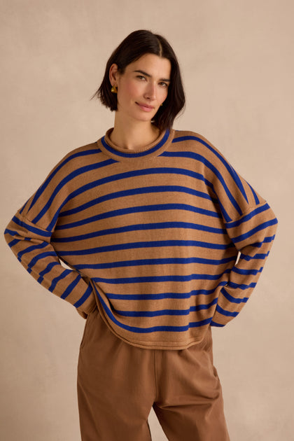 A woman with short dark hair stands with her hands on her hips against a beige background, wearing the Royal Blue Stripe Boxy Merino Knit sweater—crafted from soft merino wool in brown and blue stripes—paired with brown pants.