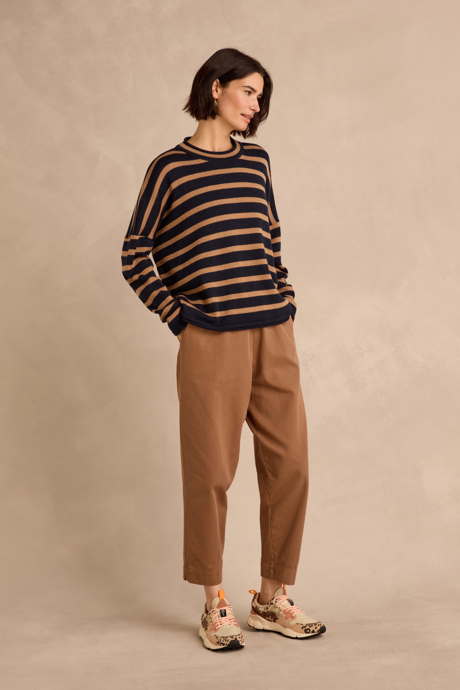 A woman stands against a beige background wearing the Navy Stripe Boxy Merino Knit sweater—crafted from soft merino wool in navy and tan stripes—paired with tan pants and floral sneakers, her hands in her pockets.