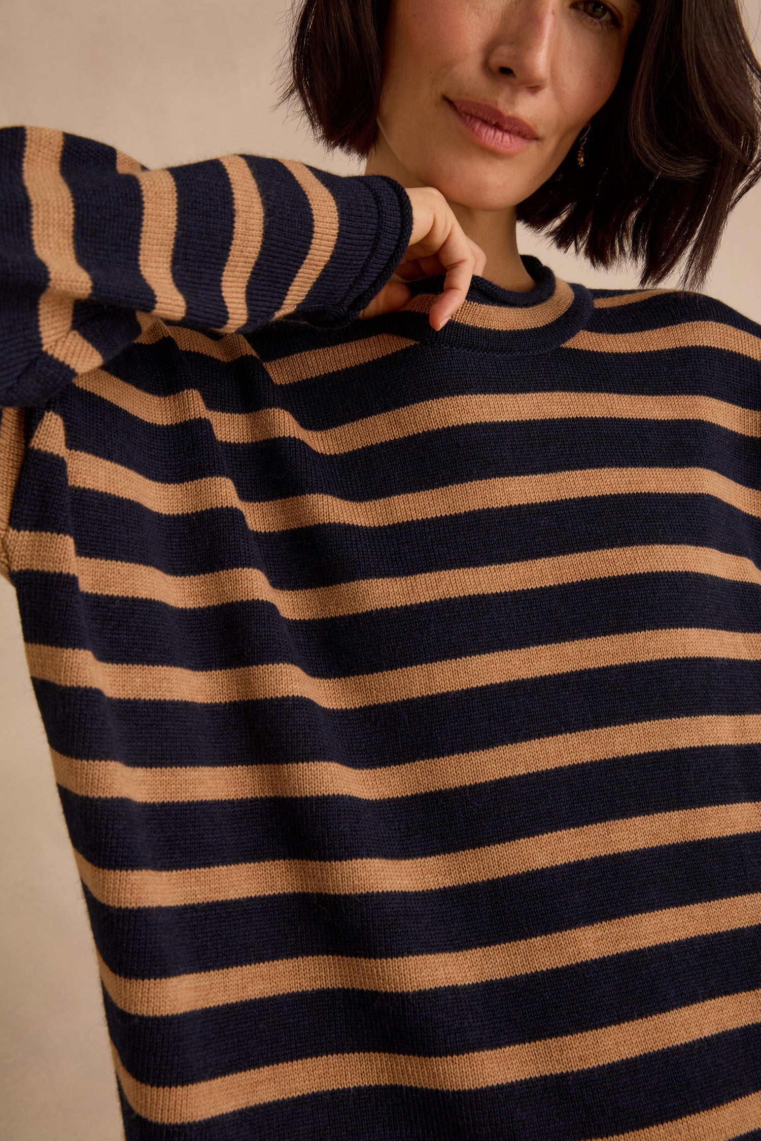 Wearing the Navy Stripe Boxy Merino Knit, a soft merino wool sweater featuring navy blue and tan stripes, a person with short dark hair—partially covering their face—raises one arm.