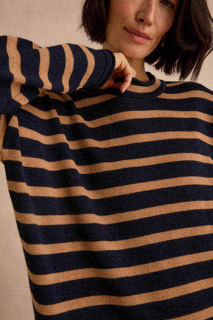 Wearing the Navy Stripe Boxy Merino Knit, a soft merino wool sweater featuring navy blue and tan stripes, a person with short dark hair—partially covering their face—raises one arm.