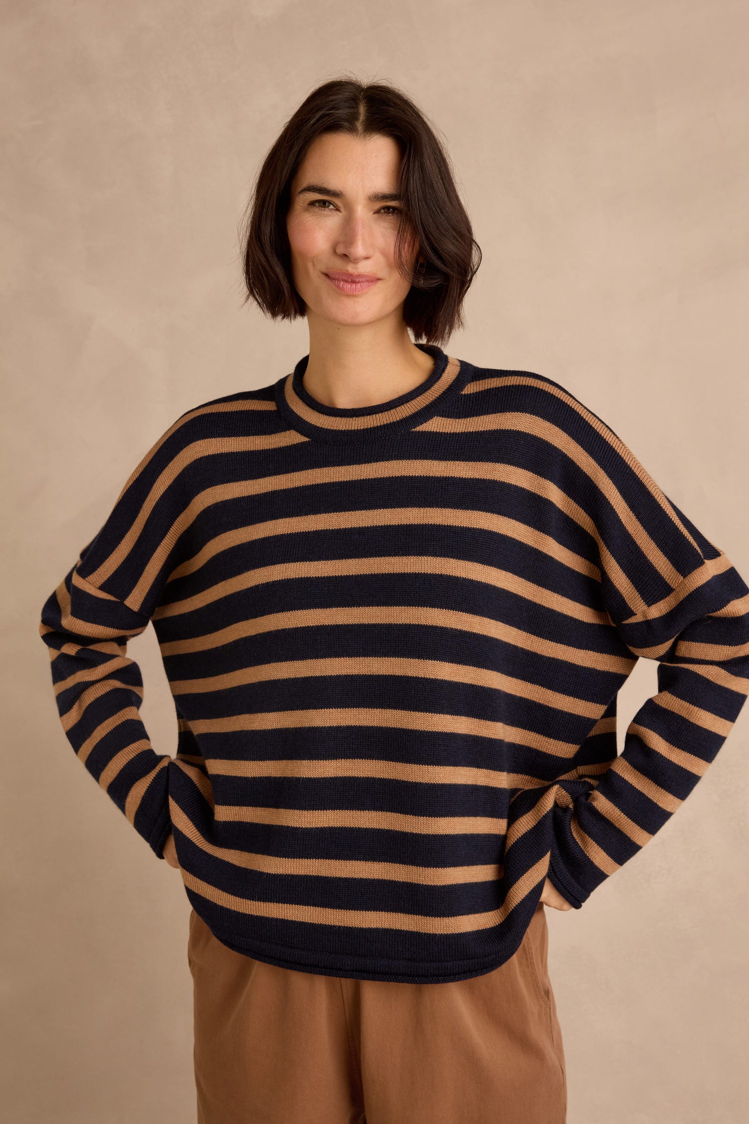 A person stands against a neutral background, wearing the Navy Stripe Boxy Merino Knit sweater and brown pants, with hands on hips and a slight smile.