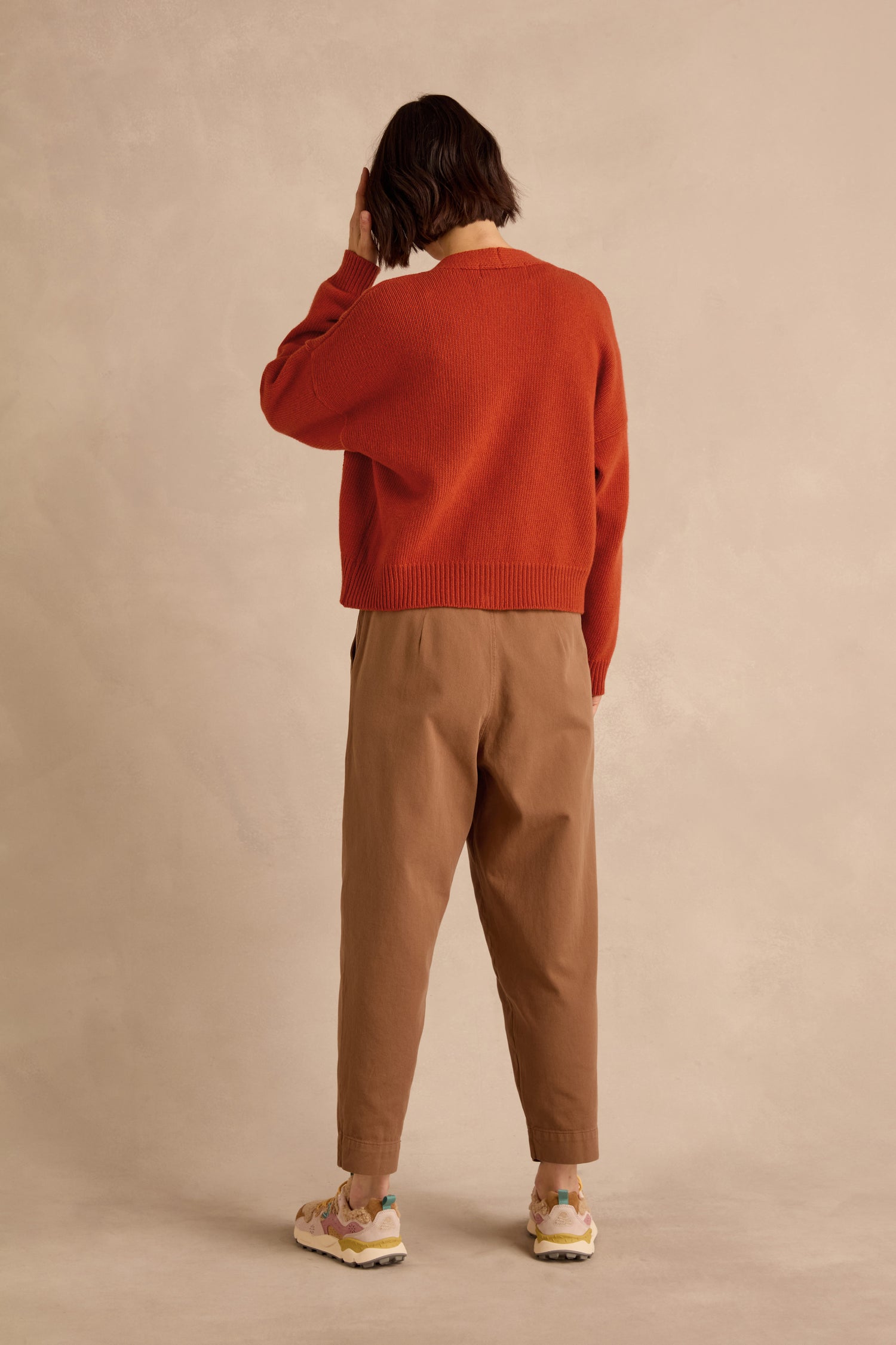 A person stands facing away, wearing the Wool Boxy Cardigan in red—a recycled wool sweater with a cropped fit—paired with tan pants and sneakers, one hand raised to their head. The background is plain beige.