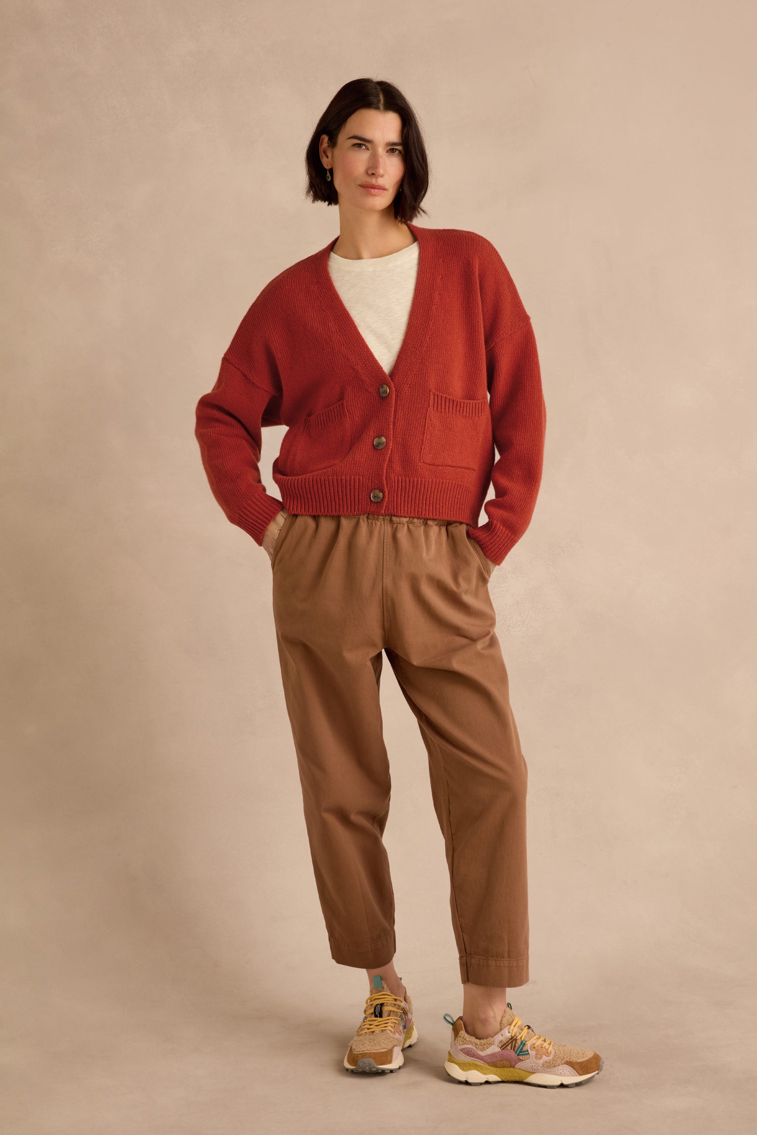 A person stands against a beige background wearing the Wool Boxy Cardigan in red—a cropped, boxy fit crafted from recycled wool—styled with a white shirt, brown pants, and yellow sneakers, hands in pockets.