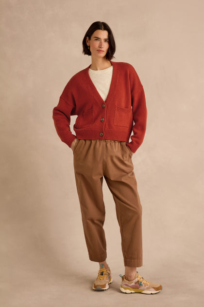 A person stands against a beige background wearing the Wool Boxy Cardigan in red—a cropped, boxy fit crafted from recycled wool—styled with a white shirt, brown pants, and yellow sneakers, hands in pockets.