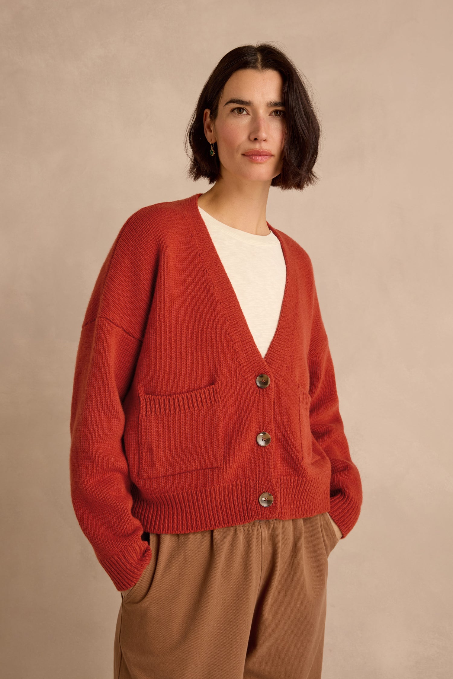 A woman with short dark hair wears the Wool Boxy Cardigan—crafted from recycled wool in a vibrant red hue—layered over a white top and brown pants, standing against a neutral background.
