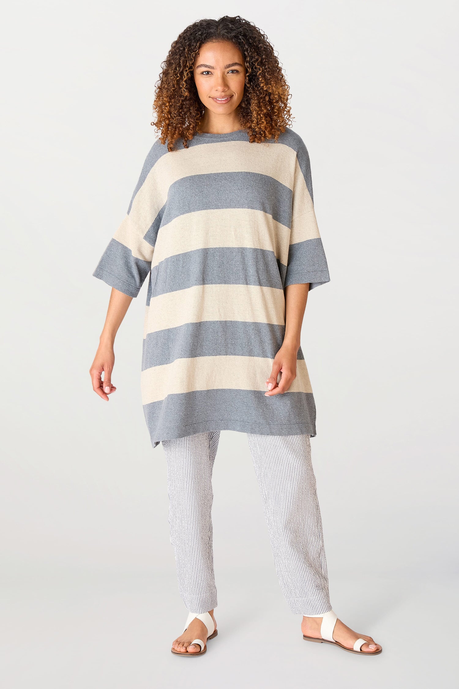 A woman in a Vera Stripe Top and textured pants made from a recycled cotton blend stands confidently, dressed in a modern, casual outfit.