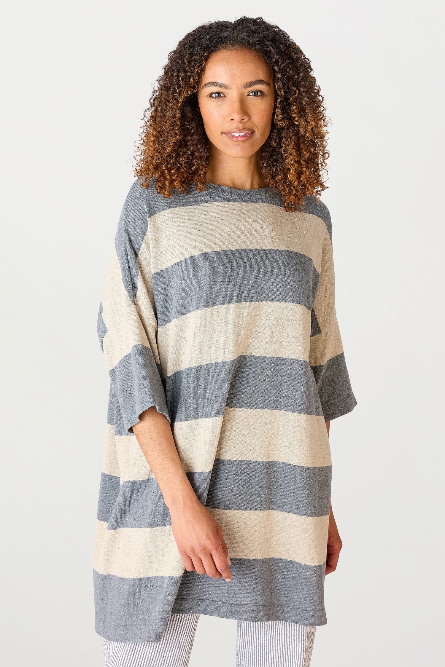 A woman with curly hair stands confidently, wearing the Vera Stripe Top made from a recycled cotton blend and striped leggings, against a plain white background.