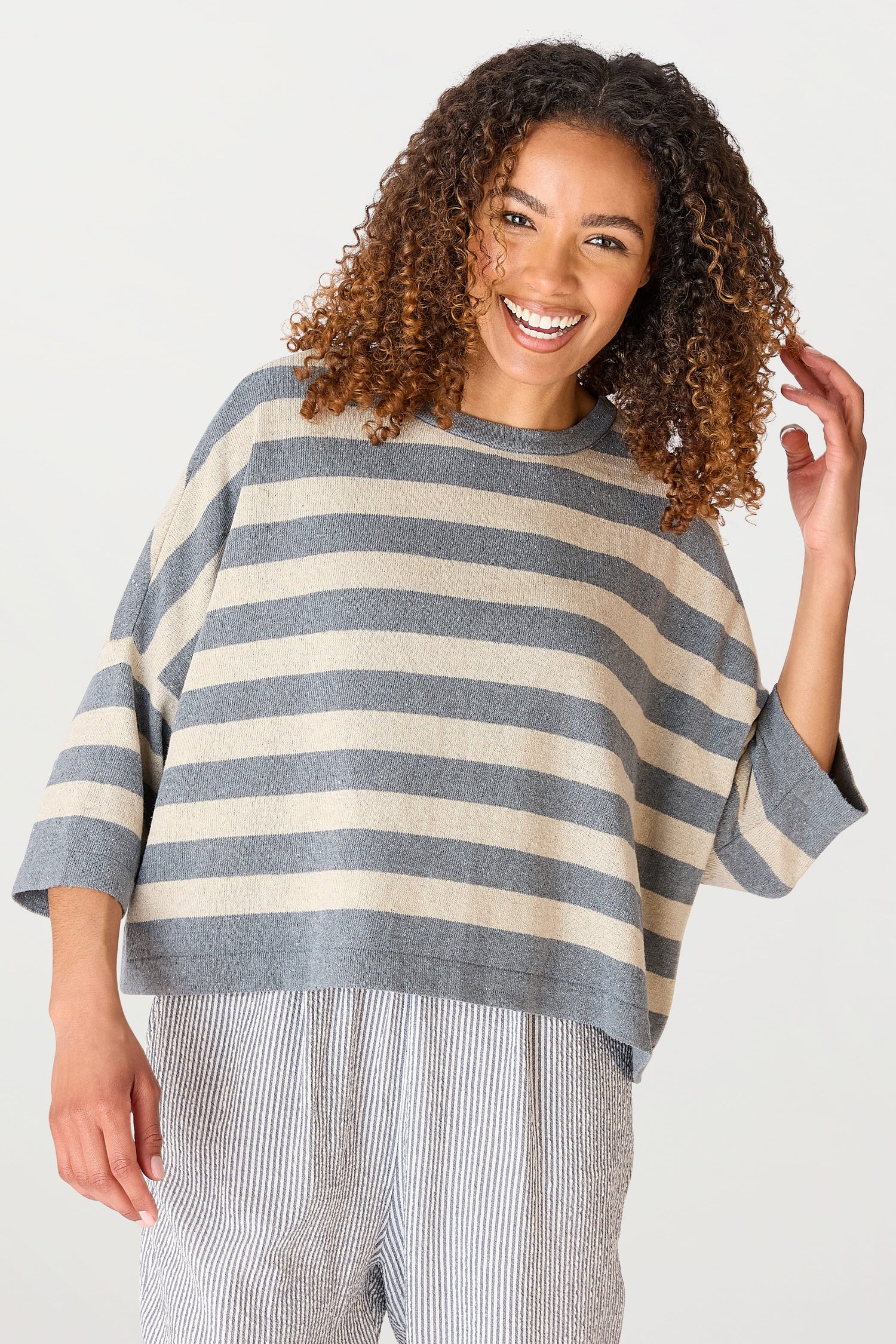 A woman with curly hair, smiling, wearing a Vibe Stripe Top and striped pants, posing against a plain background.