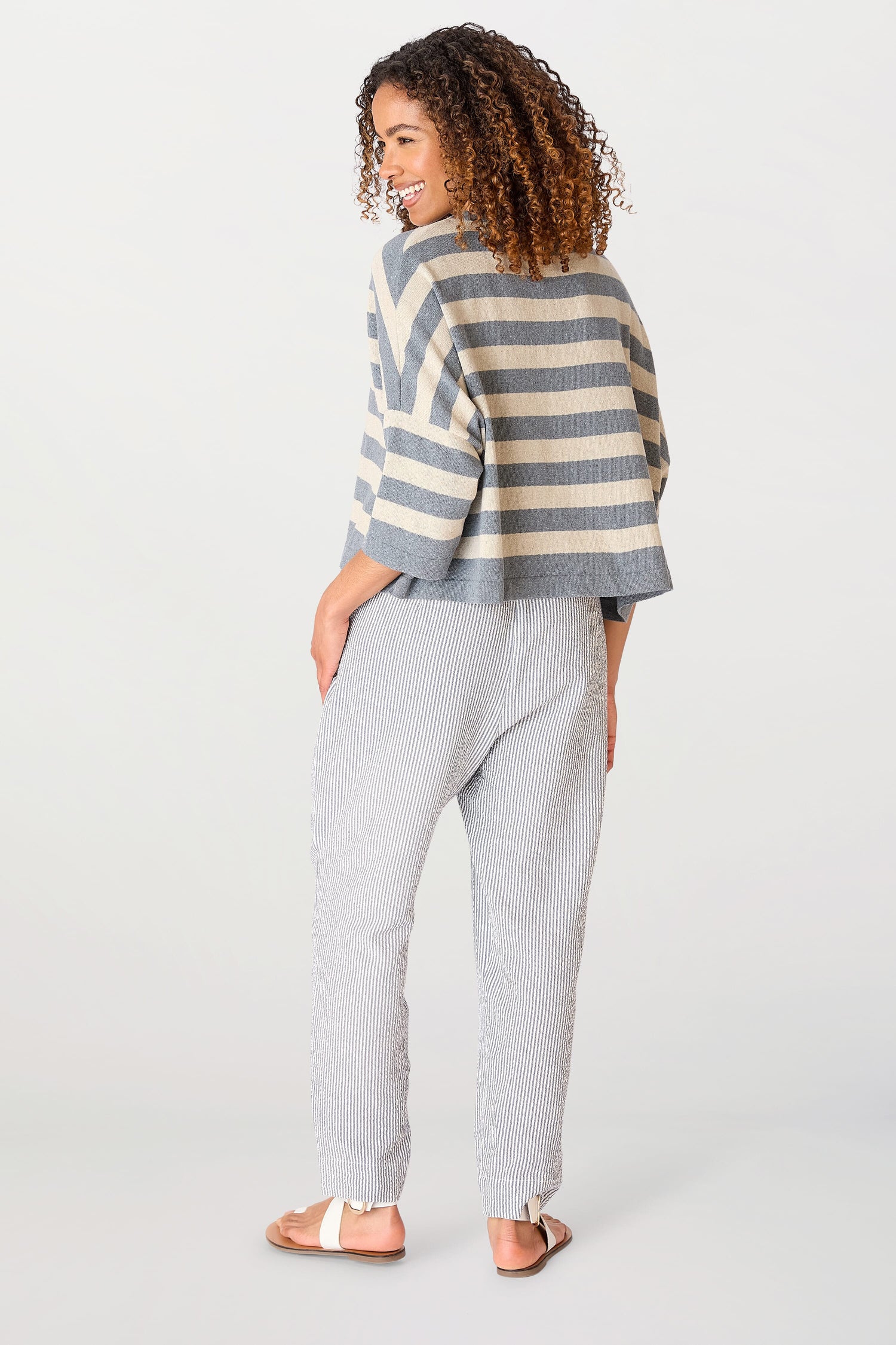 Woman standing facing away, looking over her shoulder, wearing a Vibe Stripe Top of recycled cotton, patterned pants, and sandals on a white background.