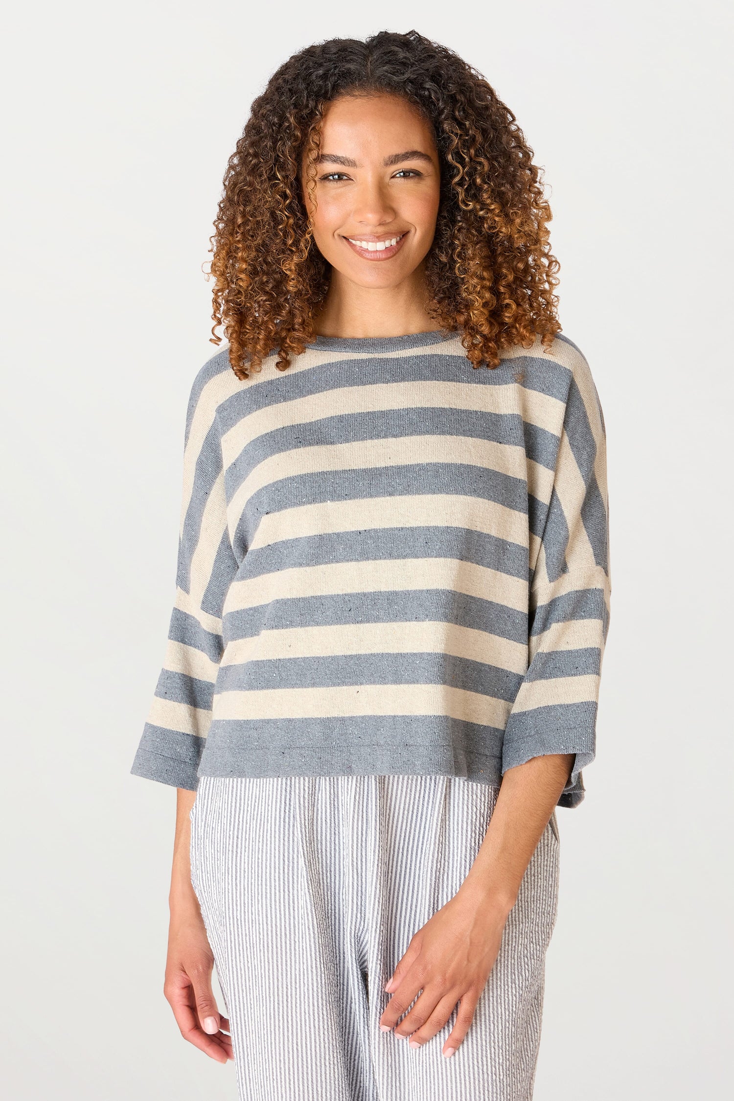 A woman with curly hair smiling, wearing a Vibe Stripe top and striped pants made of recycled cotton, standing against a white background.