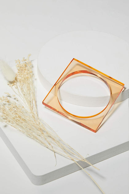 Square Bangle: a translucent orange modern accessory displayed on white geometric platforms next to dried, light-colored flowers.