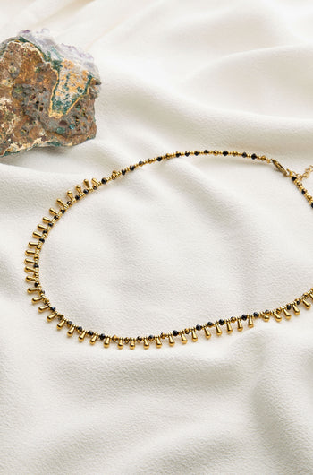 An Enyo Necklace with a gold beaded design and a series of small dangling elements by Nataraj Collections is displayed on white fabric next to a rock with green and brown hues.