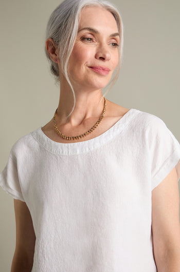 A woman with grey hair, wearing a white shirt and the Enyo Necklace from Nataraj Collections, which showcases an intricate gold beaded design, stands against a neutral background.