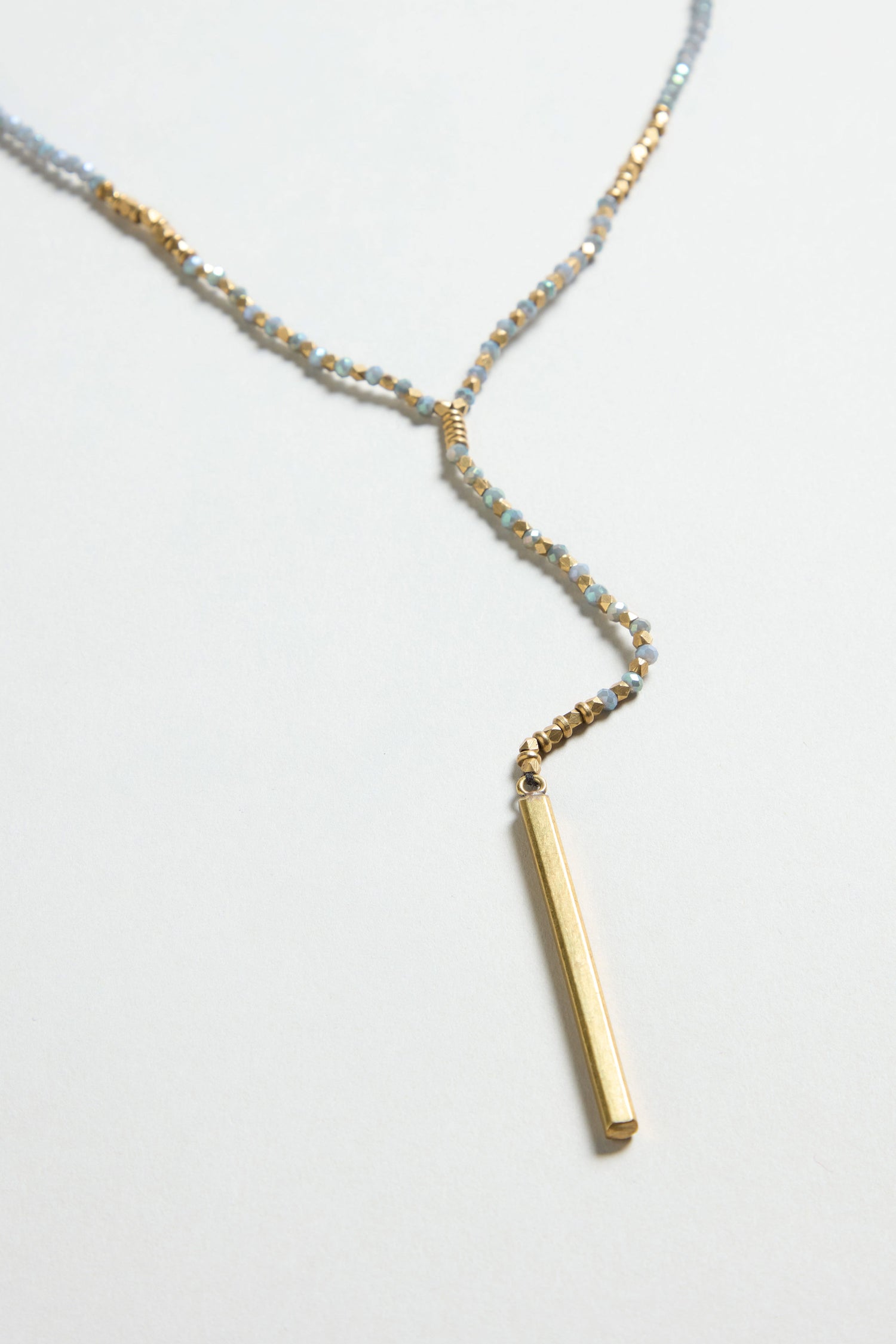 The Aditi Necklace, featuring small blue and gold beads with a vertical gold bar pendant, is displayed on a light background.