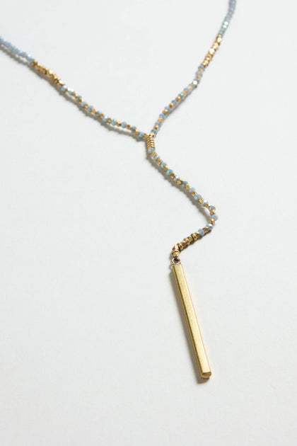 The Aditi Necklace, featuring small blue and gold beads with a vertical gold bar pendant, is displayed on a light background.