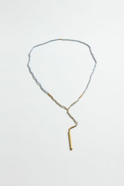 The Aditi Necklace is a delicate gold necklace with small light blue pearl beads, gold chain sections, and a vertical pendant hanging from a drop chain.