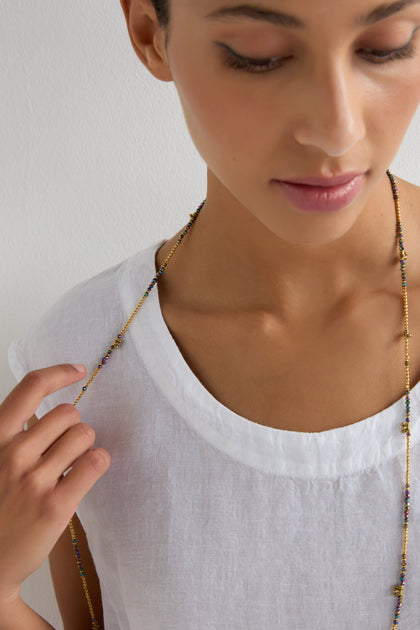 A person in a white linen top holds the Long Saros Beaded Necklace, its multicoloured seed beads from the Nataraj Collection resting near their chest as they gaze downward with a neutral expression.
