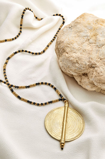 The Spiral Pendant, a long gold and black beaded necklace with a large round gold charm, is elegantly placed on white fabric near a textured beige rock.