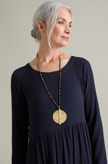 A person with gray hair wearing a dark, long-sleeved dress and a large, ornate Spiral Pendant handcrafted by skilled artisans, featuring a gold and black beaded design, stands against a plain background.