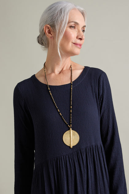 A person with gray hair wearing a dark, long-sleeved dress and a large, ornate Spiral Pendant handcrafted by skilled artisans, featuring a gold and black beaded design, stands against a plain background.