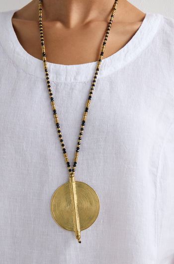 A person wearing a white linen shirt with a beaded necklace showcasing the Long Spiral Pendant in gold and black, a statement piece of artisanal craftsmanship.