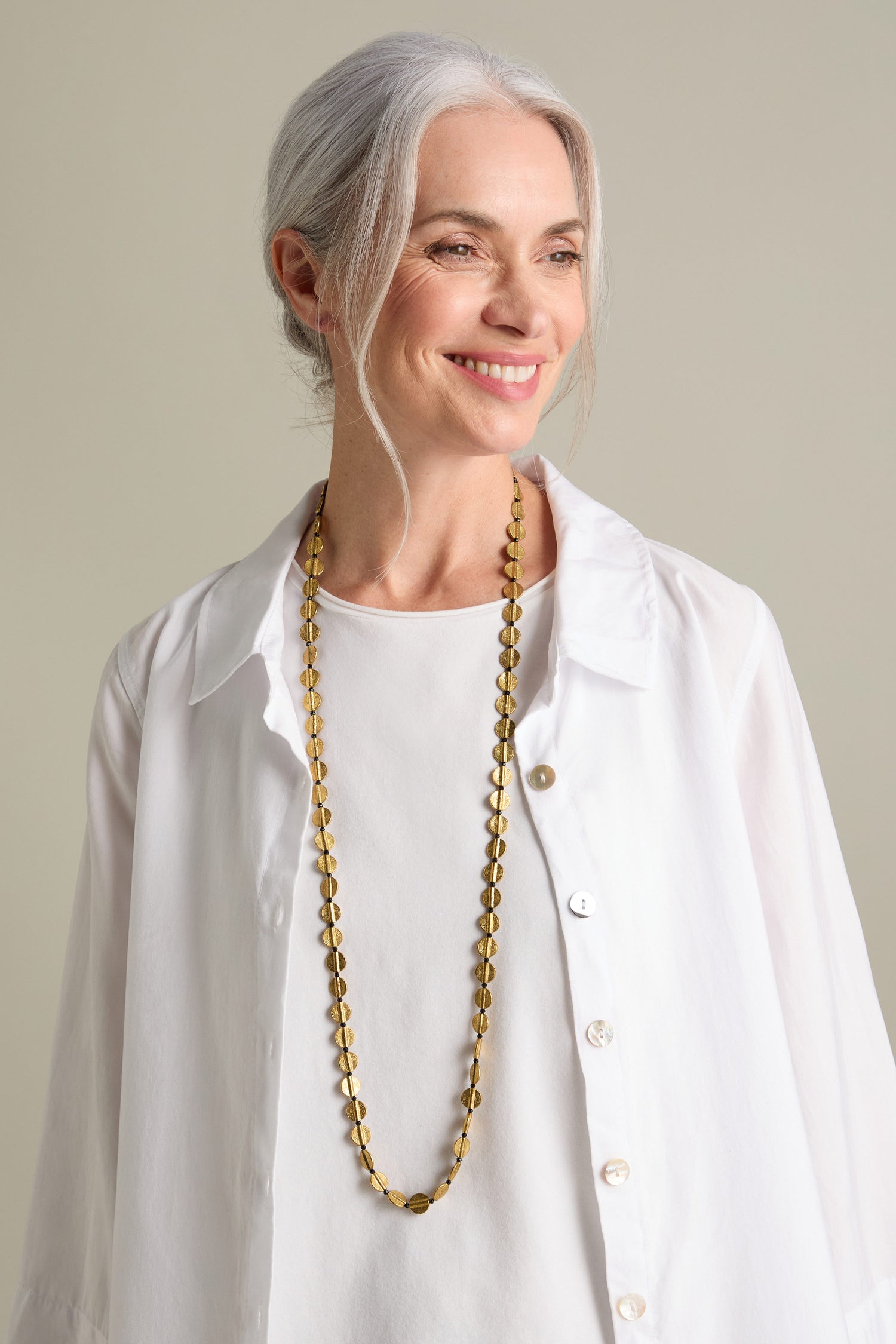 A woman with gray hair, wearing a white blouse and the Engraved Circles Necklace crafted by Jaipur artisans, smiles while looking to her right.