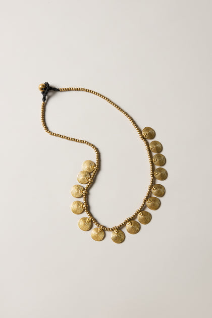 The Short Gold Chakra Necklace features golden discs with round, textured coins evenly spaced along the chain, displayed flat on a neutral background for a timeless statement look.