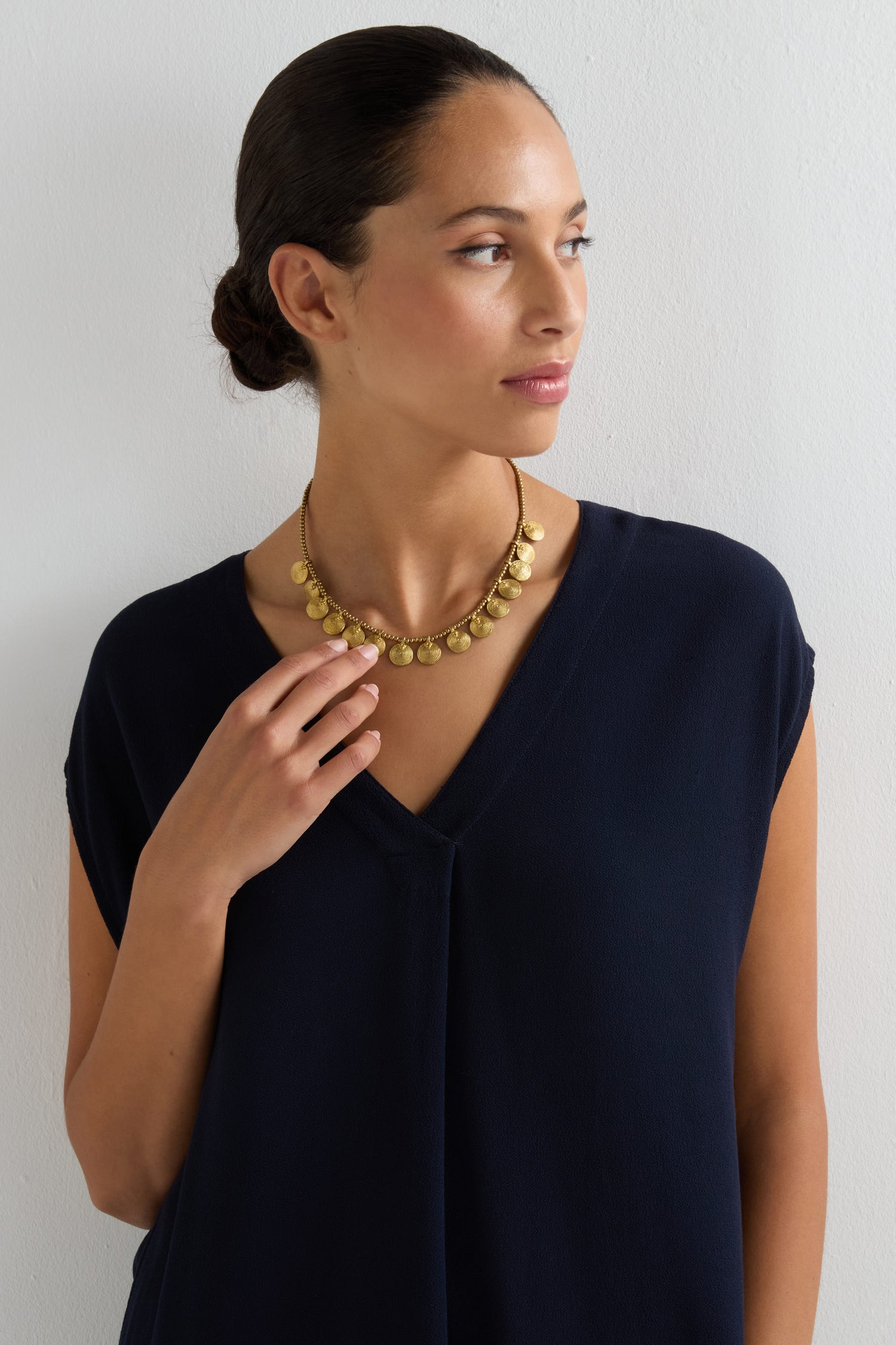 A woman in a dark blue V-neck top wears the Short Gold Chakra Necklace, featuring golden discs, as she stands against a white wall and looks to the side—a timeless statement of elegance.