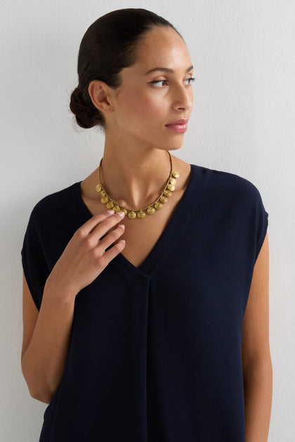 A woman in a dark blue V-neck top wears the Short Gold Chakra Necklace, featuring golden discs, as she stands against a white wall and looks to the side—a timeless statement of elegance.