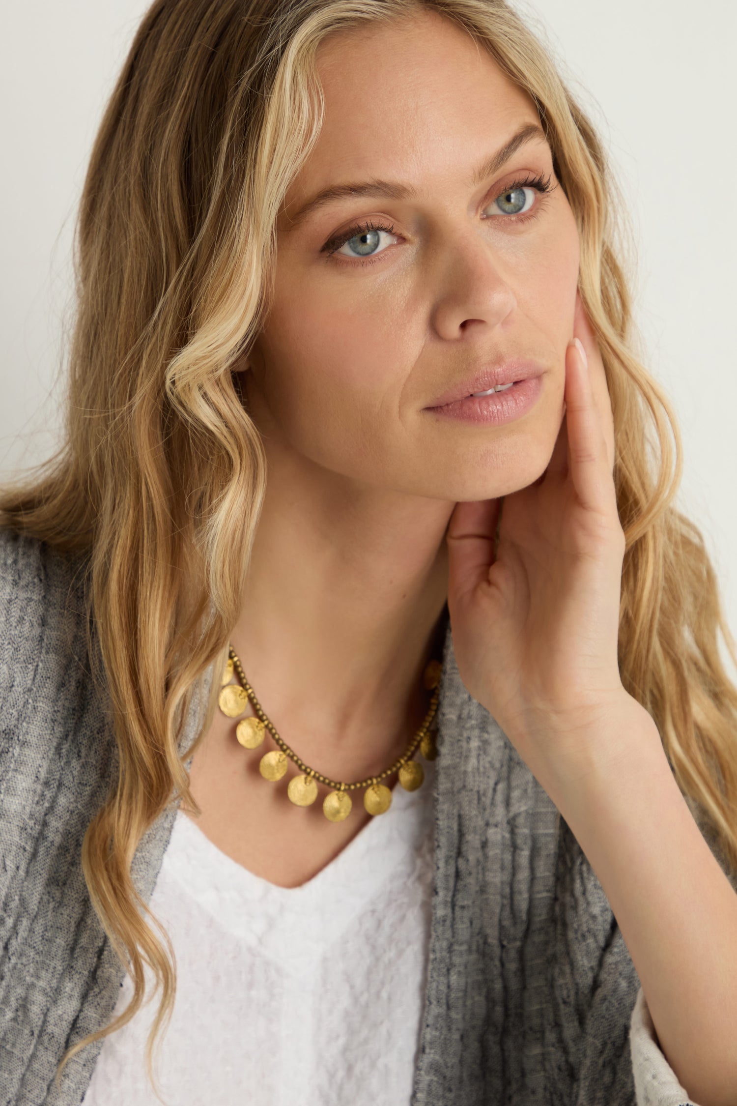 A woman with long blonde hair rests her chin on her hand, gazing slightly away from the camera, while wearing a Short Gold Chakra Necklace, a white shirt, and a grey sweater.