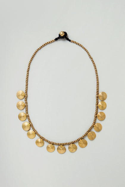 The Short Gold Chakra Necklace features small round beads and sculpted brass coin-shaped pendants, evenly spaced along the lower half, showcased on a plain light background.